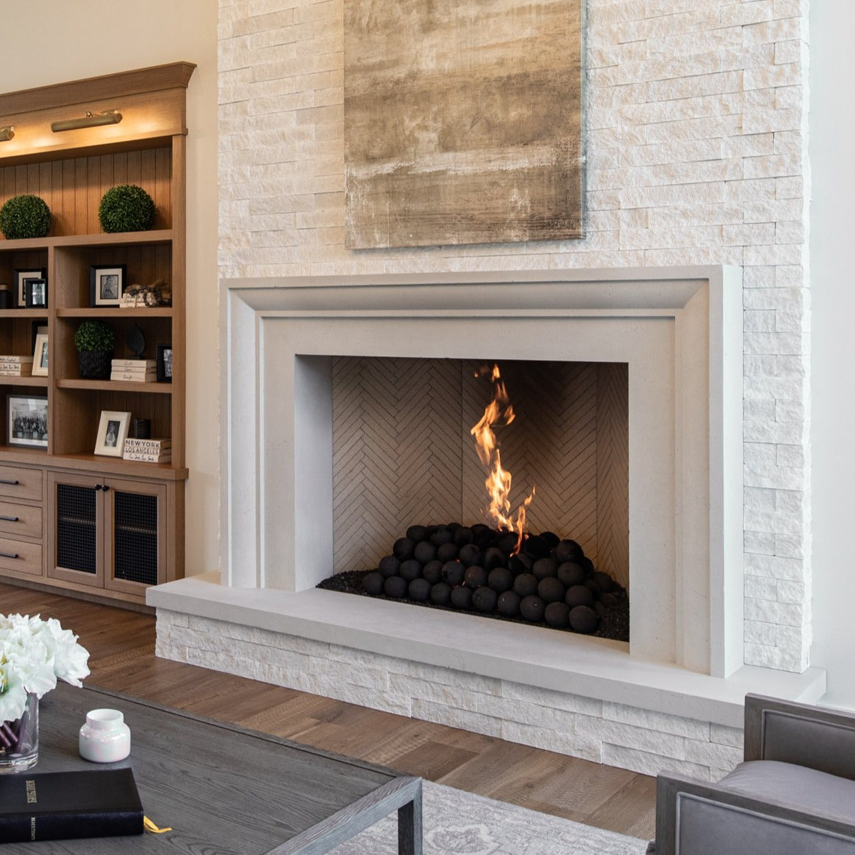 A modern white cast stone fireplace surround with a visible fire in the fireplace, situated in a well-lit room with wooden flooring and bookshelves in the background.